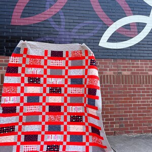 May include: A red, white, and gray patchwork quilt with a checkered pattern. The quilt is folded and leaning against a brick wall.