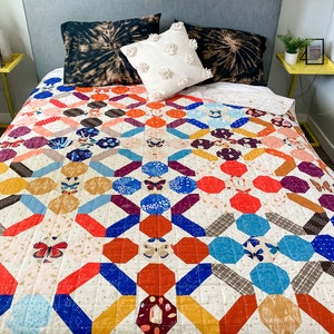 May include: A colourful quilt with a geometric pattern featuring butterflies and stars. The quilt is made with a variety of fabrics in shades of blue, red, yellow, and white. It is displayed on a bed with black and white tie-dye pillowcases.