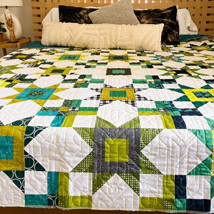 May include: A white quilt with a star pattern in shades of green, yellow, and blue. The quilt is on a bed with white pillows and a wooden headboard.