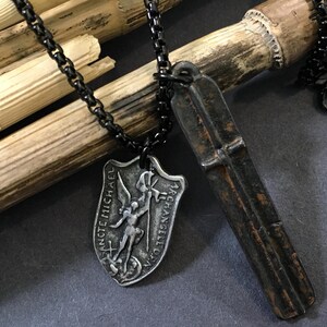 May include: Two silver necklaces with black chains. One necklace has a pendant with a depiction of an angel with the text "SANCTE MICHAEL ARCHANGELI ORA PRO NOBIS". The other necklace has a long, narrow pendant with a cross.