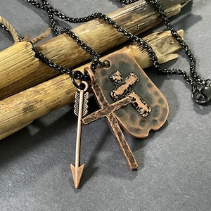 May include: A copper-toned pendant necklace featuring a cross and an arrow charm. The necklace has a black chain and a dog tag-style pendant with a cross design. The arrow charm is pointing downwards.