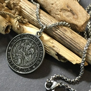 Op de afbeelding: Een zilveren ketting met een ronde hanger met een afbeelding van Sint Christoffel. De hanger heeft de tekst "Sint Christoffel Bescherm Ons".