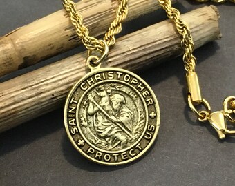 May include: Gold chain necklace with a round pendant featuring a depiction of Saint Christopher carrying a child. The pendant is inscribed with "Saint Christopher + Protect Us".