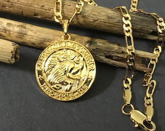 May include: A gold-colored pendant necklace featuring a round medallion with a detailed relief of a figure. The necklace has a matching gold chain with a lobster clasp. The pendant and chain are displayed on a dark surface.