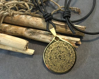 May include: A gold pendant necklace with a circular design featuring a detailed Aztec calendar. The necklace is on a black leather cord.