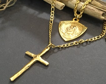 May include: A gold chain necklace with two pendants. One pendant is a shield-shaped charm with the text "SAN CRISTOBAL" and a religious image. The other pendant is a hammered gold cross.
