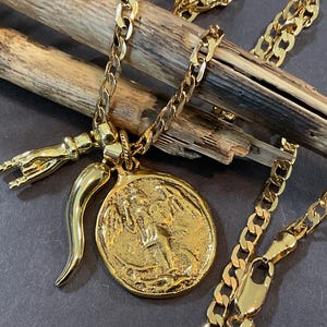 May include: A gold-toned metal chain necklace featuring three charms: a circular coin-style pendant with an embossed figure, a smooth curved horn-shaped charm, and a small hand charm displaying a gesture with extended index and pinky fingers.
