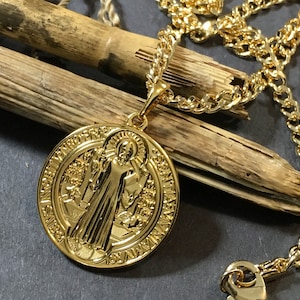 May include: A gold-colored pendant necklace featuring a detailed medallion with a religious figure and Latin inscription. The pendant hangs from a curb chain. The necklace rests on a wooden surface.