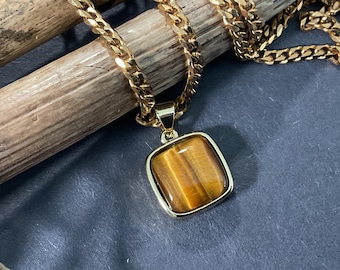 May include: A gold-toned necklace with a square pendant featuring a tiger's eye stone. The chain is a thick curb link style. The pendant is framed in a gold setting. The necklace rests on a dark blue surface.