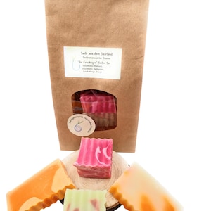 Könnte beinhalten: Ein Set aus vier handgefertigten Seifen in einer braunen Papiertüte. Die Seifen sind in verschiedenen Farben und Mustern, darunter Orange, Pink und Grün. Die Tüte hat ein Etikett mit der Aufschrift "Seife aus dem Saarland" und "die Fruchtigen Seifen Set".