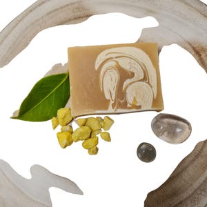 May include: A bar of handmade soap with a swirl pattern in shades of brown and white. The soap is sitting on a light brown surface with small yellow stones and clear glass stones.