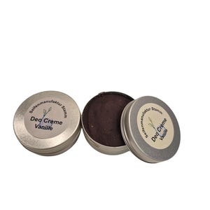 Op de afbeelding: Drie kleine, zilverkleurige blikjes deodorantcrème. Eén blikje is open en toont een donkerbruine, vaste crème. De deksels zijn voorzien van de tekst "Deo Creme Vanille" met een bladmotief. Het product is van "Seifenmanufaktur Stamm".