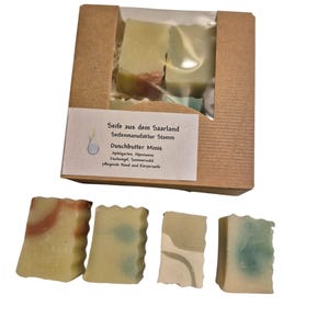 Peut inclure: Un ensemble de quatre petits savons rectangulaires dans une boîte en carton. Les savons sont de différentes nuances de vert et de brun. La boîte porte une étiquette qui indique "Seife aus dem Saarland Seifenmanufaktur Stamm Duschbutter Minis Apfelgarten, Alpenwiese Dschungel, Semmerwald pflegende Hand und Körperseife".