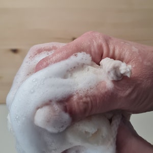 May include: A hand squeezing a white, foamy bath pouf. The pouf is covered in white bubbles and is being squeezed in a cupped hand.