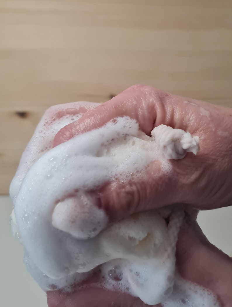 May include: A hand squeezing a white, foamy bath pouf. The pouf is covered in white bubbles and is being squeezed in a cupped hand.