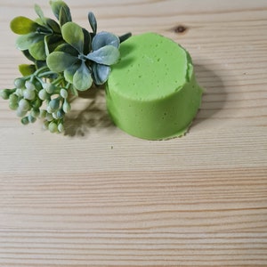 Könnte beinhalten: Eine runde, grüne, feste Seife auf einer Holzoberfläche mit einer kleinen grünen Pflanze.