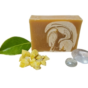 May include: A bar of handmade soap with a swirl design in shades of beige and white. The soap is surrounded by small yellow pieces of dried fruit and clear stones.