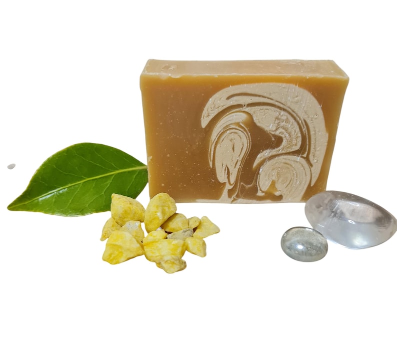 May include: A bar of handmade soap with a swirl design in shades of beige and white. The soap is surrounded by small yellow pieces of dried fruit and clear stones.