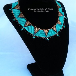 May include: A turquoise and gold beaded necklace with a geometric design. The necklace is on a black velvet stand. "Designed by Deborah Finkle for Shoshin Arts" is written on the stand.