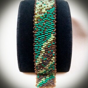 May include: A green, brown, and gold beaded bracelet with a geometric pattern. The bracelet is on a black display stand.