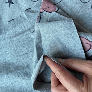 Op de afbeelding: Close-up van grijze stof met een patroon van sterren en cartoongezichten. De stof is gevouwen en toont de achterkant. De stof heeft een zachte, getextureerde uitstraling, geschikt voor kleding of woondecoratie.
