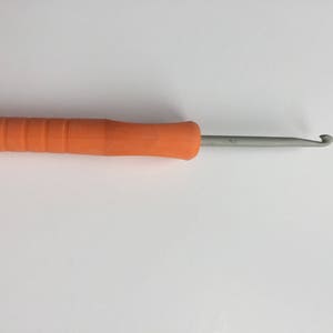 Puede incluir: Un gancho de ganchillo naranja con un gancho de metal. El gancho mide aproximadamente 20 cm de largo.
