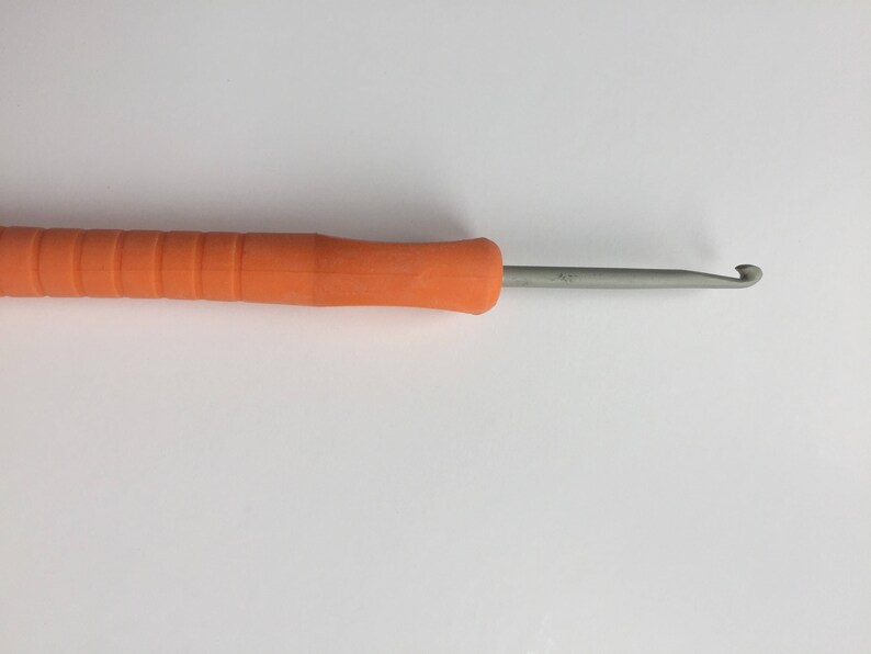 Puede incluir: Un gancho de ganchillo naranja con un gancho de metal. El gancho mide aproximadamente 20 cm de largo.