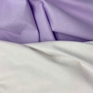 Puede incluir: Primer plano de dos telas, una en color lavanda suave y otra en blanco. Las telas est&aacute;n dobladas y drapeadas, creando sombras y texturas. La imagen resalta el contraste entre los dos colores y la suavidad de los materiales.