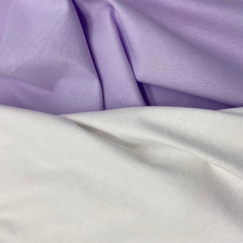 Puede incluir: Primer plano de dos telas, una en color lavanda suave y otra en blanco. Las telas est&aacute;n dobladas y drapeadas, creando sombras y texturas. La imagen resalta el contraste entre los dos colores y la suavidad de los materiales.