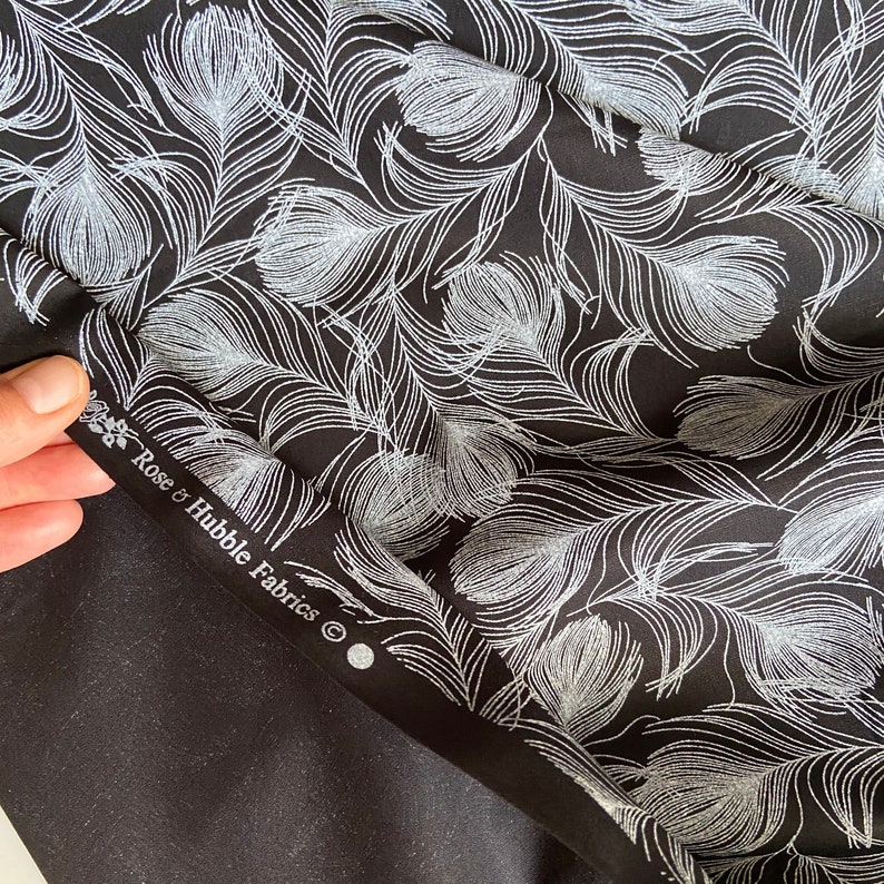 May include: Black fabric with a silver glitter feather print. The fabric has a label that reads "Rose & Hubble Fabrics".