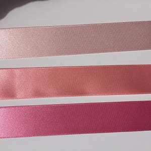 May include: Three satin ribbon spools in different shades of pink. The top spool is a light pink, the middle spool is a peach color, and the bottom spool is a deep pink.