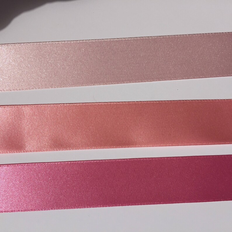 May include: Three satin ribbon spools in different shades of pink. The top spool is a light pink, the middle spool is a peach color, and the bottom spool is a deep pink.