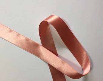 May include: A length of peach-colored satin ribbon, looped and lying on a white surface. The ribbon has a smooth, shiny texture and a subtle sheen. The edges are neatly finished with a fine stitch. Ideal for crafts and gift wrapping.