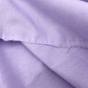 Puede incluir: Primer plano de una tela suave de color lavanda con un borde crudo e inacabado. La tela est&aacute; doblada, creando sombras y textura. El material parece ser un algod&oacute;n ligero o un textil similar.