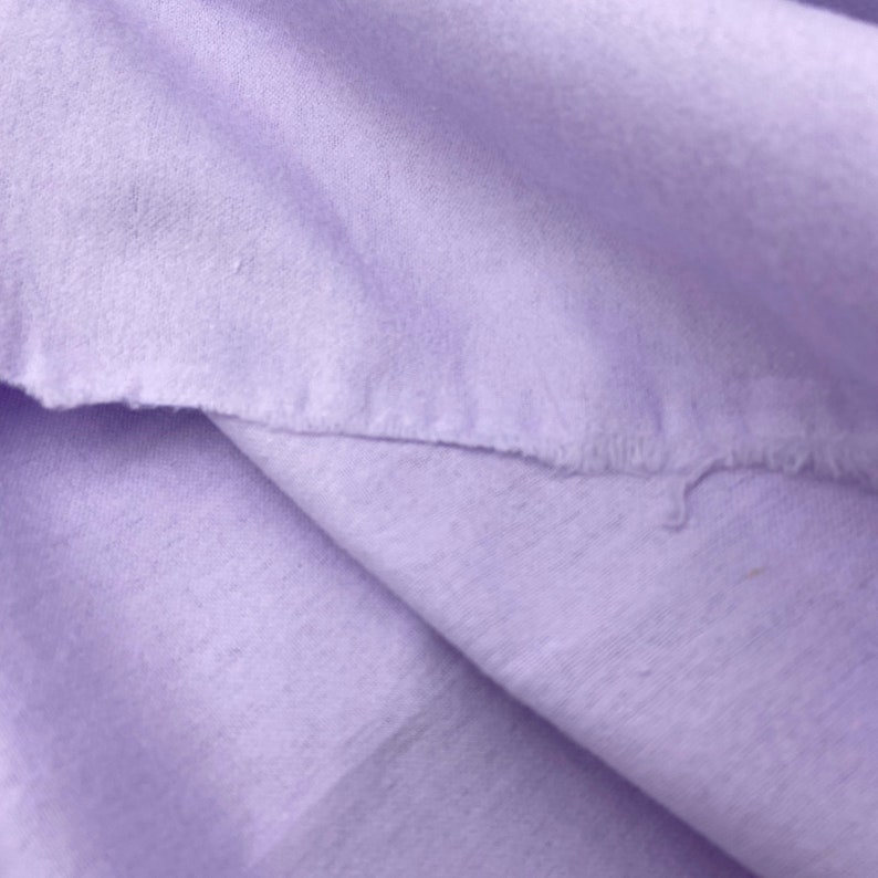 Puede incluir: Primer plano de una tela suave de color lavanda con un borde crudo e inacabado. La tela est&aacute; doblada, creando sombras y textura. El material parece ser un algod&oacute;n ligero o un textil similar.