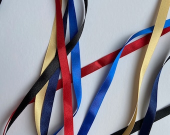 May include: Five different colored satin ribbons, including black, navy blue, red, gold, and light blue, are arranged in a crisscross pattern on a white background.
