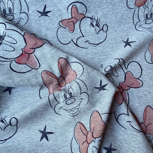 Op de afbeelding: Grijze stof met een herhaald patroon van Minnie Mouse-gezichten, roze strikken en zwarte sterren. Het gezicht van het stripfiguur is zwart omlijnd, met een roze strik en een glimlachende uitdrukking. De stof heeft een zacht, getextureerd uiterlijk.