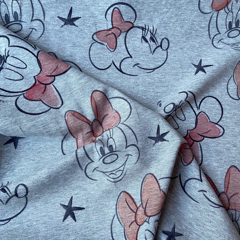 Op de afbeelding: Grijze stof met een herhaald patroon van Minnie Mouse-gezichten, roze strikken en zwarte sterren. Het gezicht van het stripfiguur is zwart omlijnd, met een roze strik en een glimlachende uitdrukking. De stof heeft een zacht, getextureerd uiterlijk.
