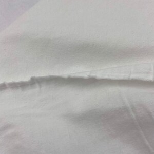 Puede incluir: Primer plano de tela blanca con borde sin rematar. La tela parece ser un material suave y tejido, posiblemente algod&oacute;n o lino. La imagen muestra la textura y la ca&iacute;da de la tela.