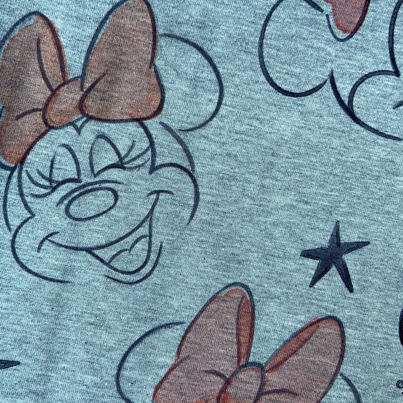 Op de afbeelding: Close-up van grijze stof met een herhaald patroon van Minnie Mouses gezicht, strik en een ster. De Minnie Mouse-gezichten zijn zwart omlijnd, met een glimlachende uitdrukking. De strikken zijn bruin.