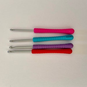 Könnte beinhalten: Vier Häkelnadeln mit verschiedenen Grifffarben: rosa, blau, lila und rot. Die Nadeln sind vertikal angeordnet, wobei die rosa Nadel oben und die rote Nadel unten ist.