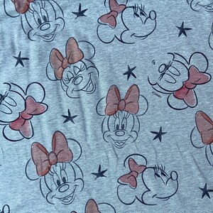 Op de afbeelding: Grijze stof met een herhaald patroon van Minnie Mouse-gezichten, roze strikken en zwarte sterren. De Minnie Mouse-gezichten zijn zwart omlijnd, met roze strikken. Het Disney-logo is zichtbaar.