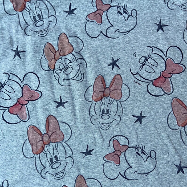 Op de afbeelding: Grijze stof met een herhaald patroon van Minnie Mouse-gezichten, roze strikken en zwarte sterren. De Minnie Mouse-gezichten zijn zwart omlijnd, met roze strikken. Het Disney-logo is zichtbaar.
