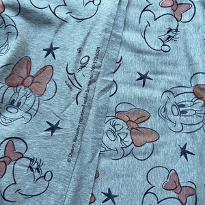 Op de afbeelding: Grijze stof met een herhaald patroon van Minnie Mouse-gezichten, rode strikken en zwarte sterren. Het ontwerp bevat de tekst "Disney. Exclusive registered under license no. 100-1754569".