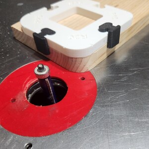 May include: A woodworking router setup. A white and black jig is clamped to a wooden board. The router bit is visible inside a red base plate, ready for use on a metal surface.