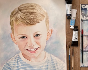 Puede incluir: Retrato en acuarela de un niño sonriente con cabello rubio y ojos azules, vistiendo una camisa a rayas azules y blancas. Suministros de arte, incluyendo tubos de pintura, pinceles y una paleta, rodean el retrato sobre una superficie de madera.