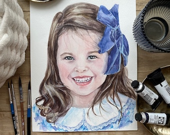 Puede incluir: Pintura de acuarela de una niña con cabello castaño que lleva un vestido azul con cuello blanco y un lazo azul en el pelo. La pintura está sobre papel blanco y está rodeada de materiales de arte.