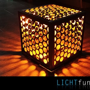 以下が含まれることがあります： 側面に円形の模様がくり抜かれた黒い立方体のランプ。ランプは点灯しており、木製の床に温かい光を投げかけています。画像の右下隅には「LICHTfunken」というテキストが表示されています。