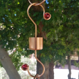 May include: Copper rain chain with red glass beads. The chain is made of copper wire and has a copper tube at the top. The beads are red and have a shiny finish.