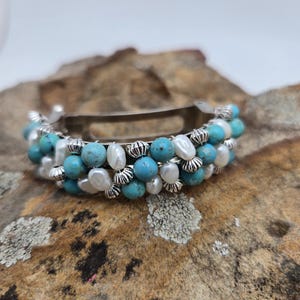 May include: A silver barrette adorned with turquoise and white pearl beads. The beads are arranged in rows, creating a layered effect. The barrette is set against a textured, brown surface.